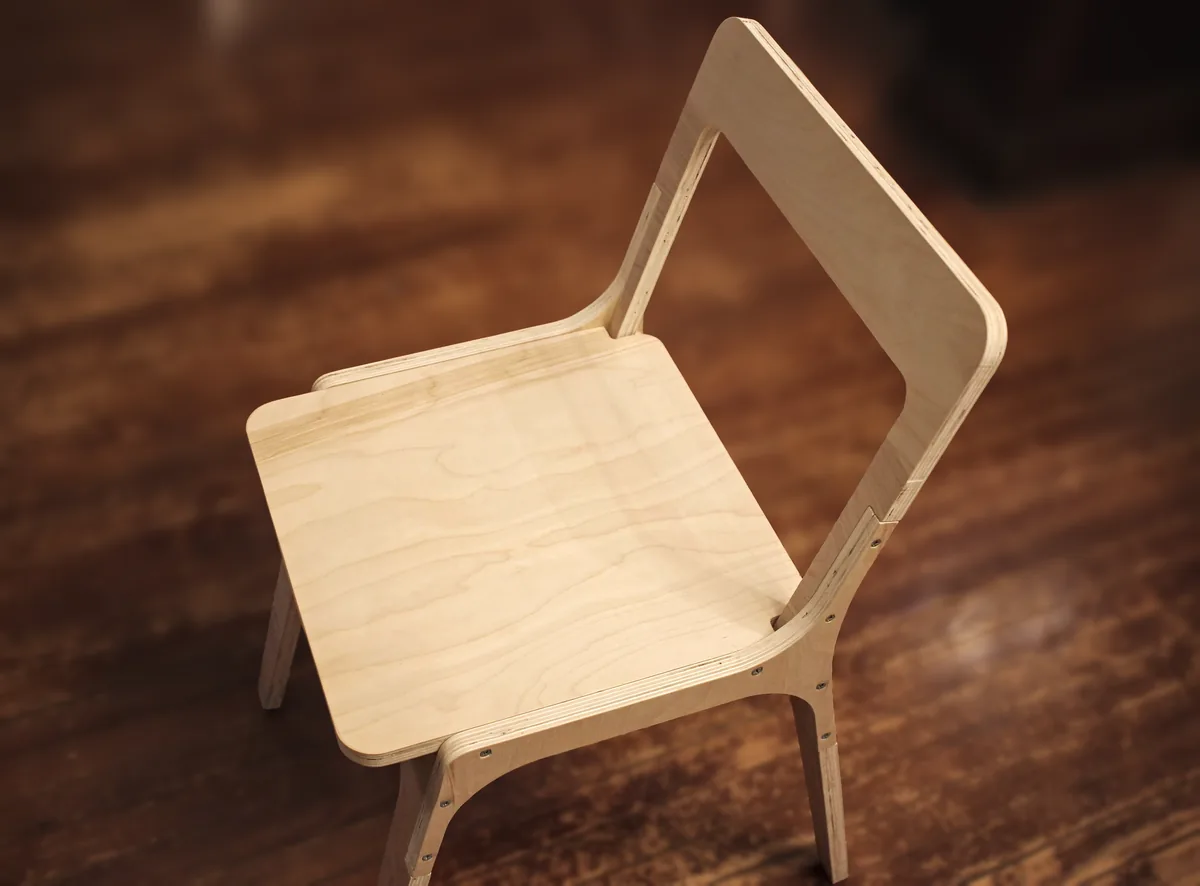A plywood chair with visible wood grain stands on a dark wood floor.