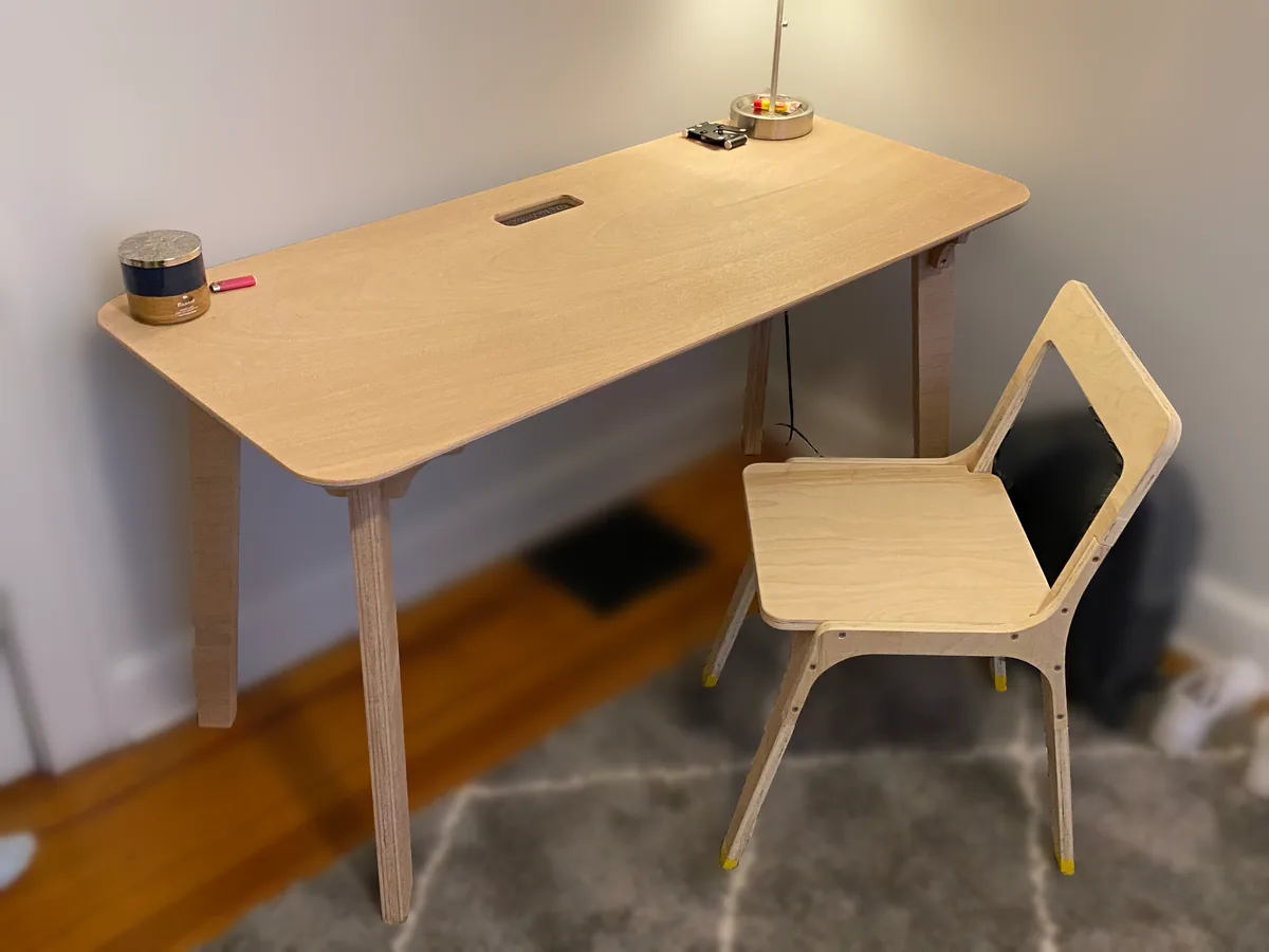 A plywood desk and matching chair stand in a room with a softly blurred background.