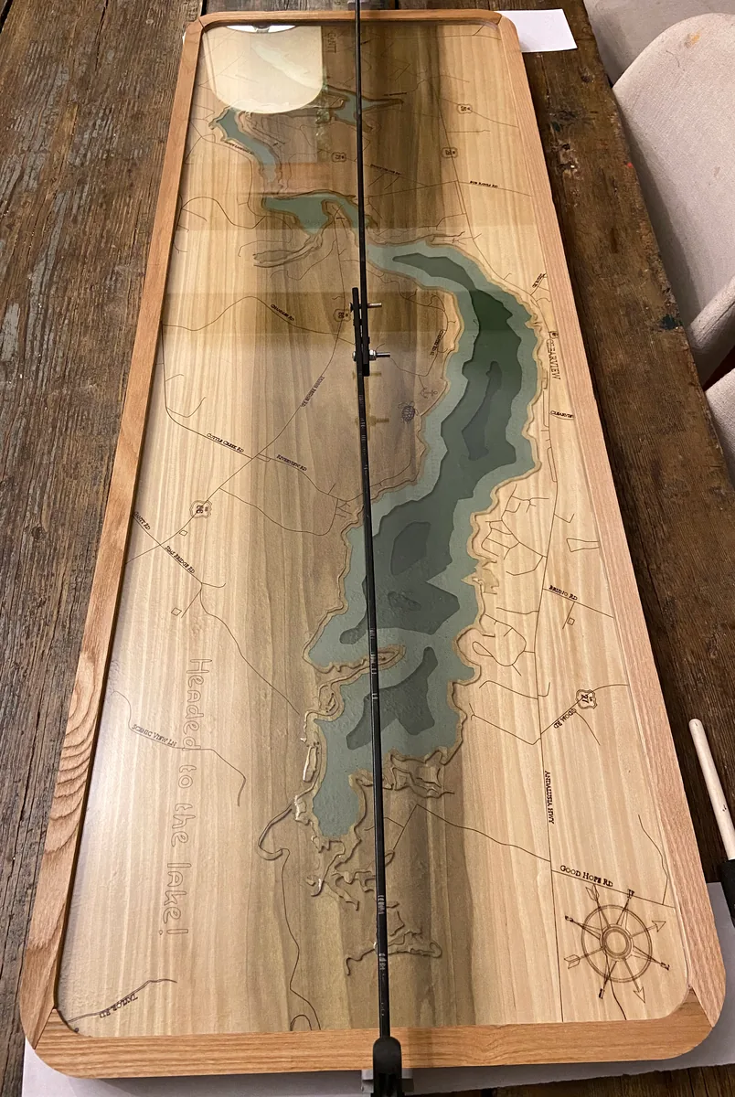 A wooden lake-map panel with green water contours rests on a table beneath a fishing rod.